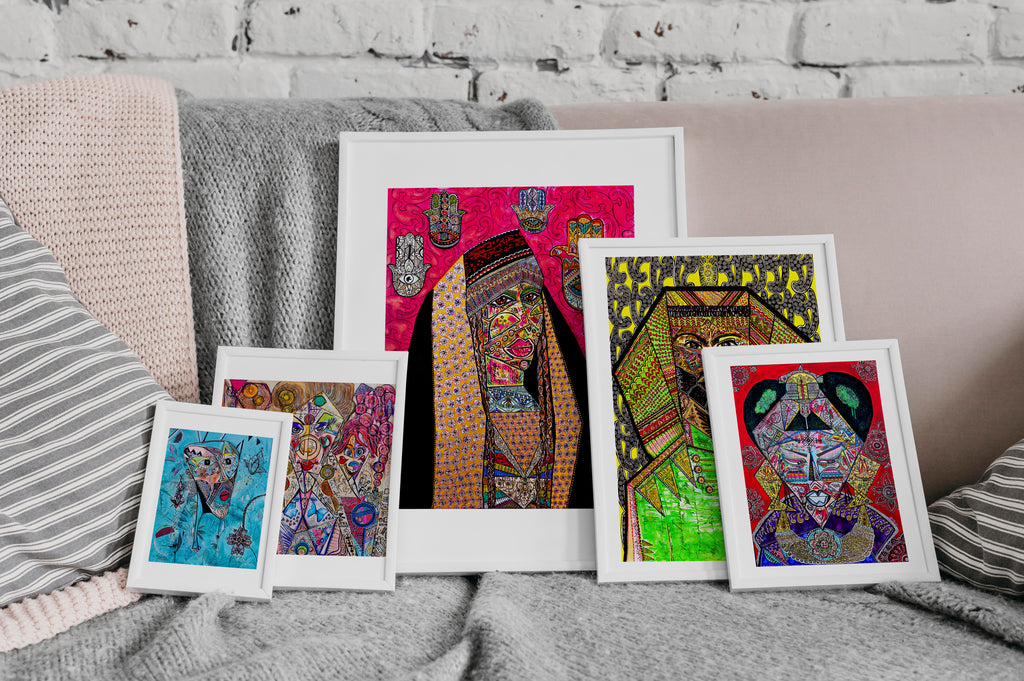 A group of Shabsoug Art prints in white frames propped together on a bed with knitted throws